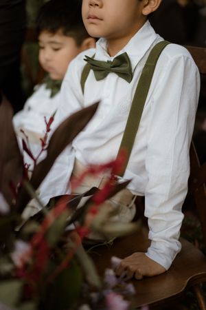 Children in white shirts with suspenders sit in a row at a formal event with red flowers in the foreground.