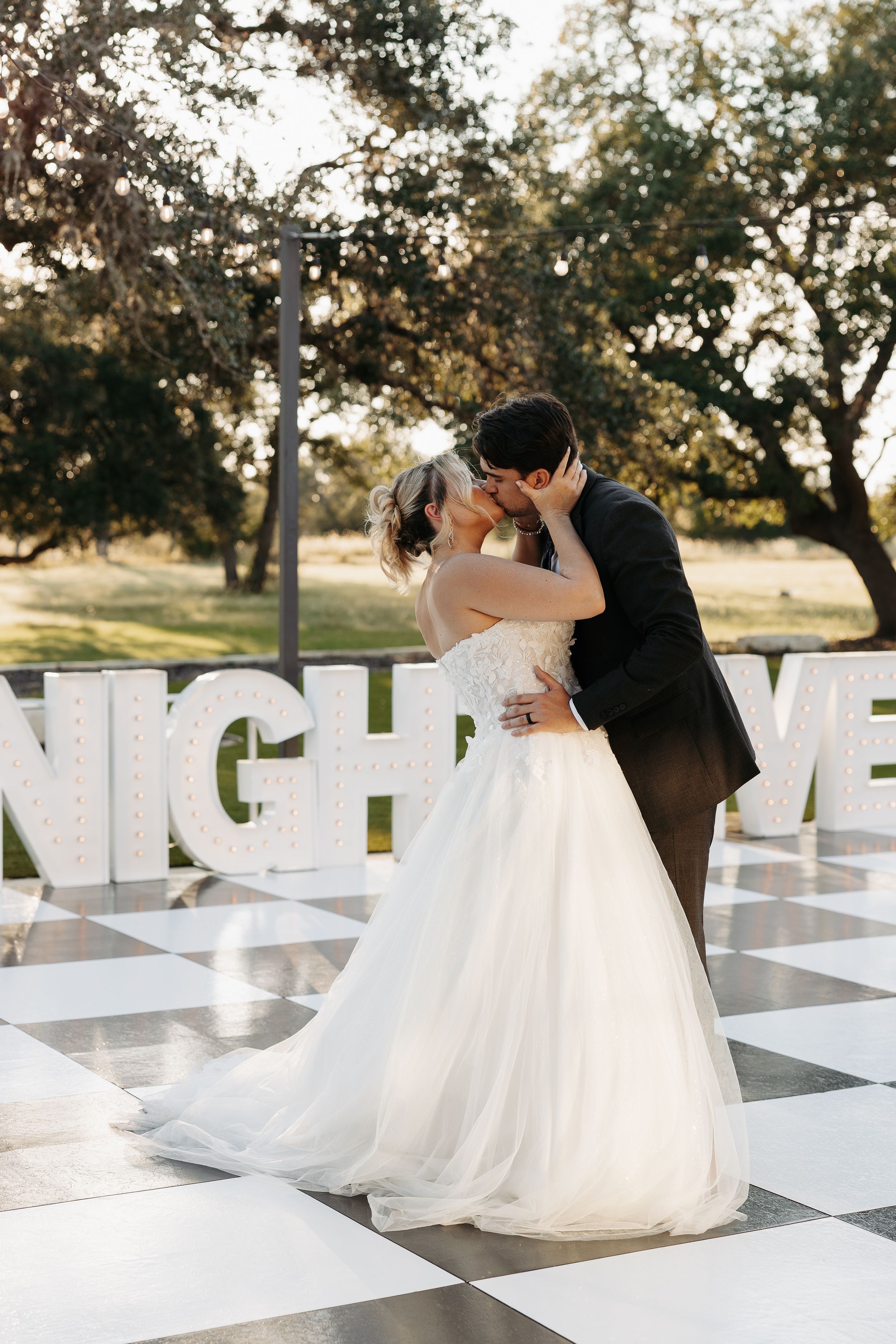 Wedding couple sharing romantic moments on a giant outdoor chess board with white chairs and trees.