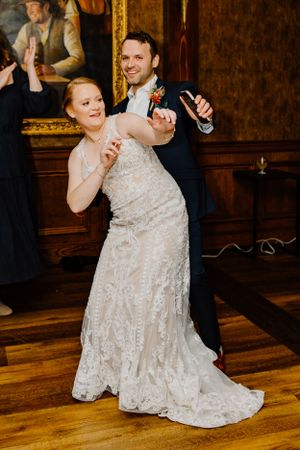 Bride in lace gown and groom dance playfully on hardwood floors during wedding reception.