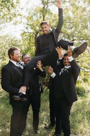 Groomsmen in black suits perform a playful lifting pose sequence in an outdoor setting with trees in the background.