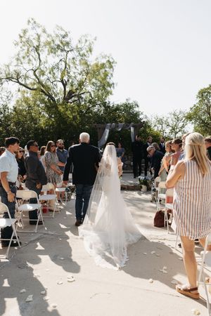 Outdoor wedding ceremony with guests seated in white chairs as the bride walks down the aisle on a sunny day.