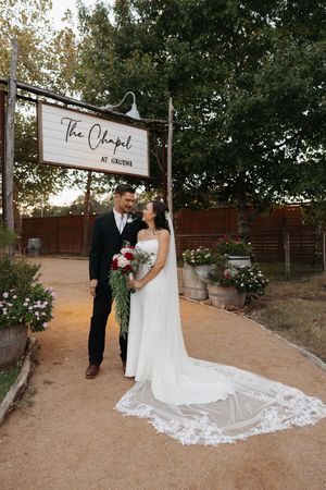 A couple in wedding attire poses together on a rustic outdoor path surrounded by trees and rose bushes.