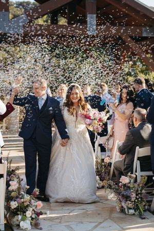 A joyful wedding couple walks down the aisle while guests shower them with confetti under a rustic outdoor pavilion.