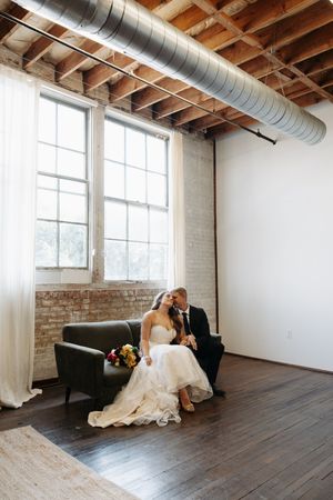 A wedding couple relaxes on a black leather couch in an industrial loft space with exposed brick walls and wooden beams.