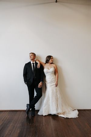 Newlywed couple poses against white wall in classic black suit and flowing white wedding gown on wooden floor.