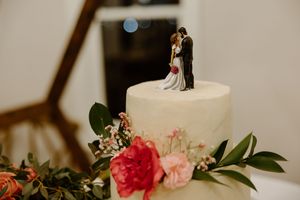 Close-up of white wedding cake decorated with traditional bride and groom topper, pink roses and green foliage.