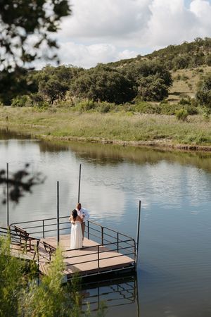 A wooden dock extends over a tranquil lake surrounded by green hills and trees on a sunny day.