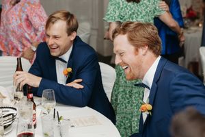 Two wedding guests in navy suits share a candid laughing moment at a reception table.