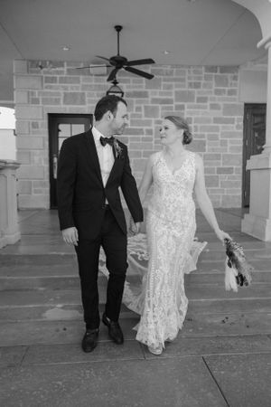 Bride in lace dress and groom in tuxedo walk together under covered walkway.