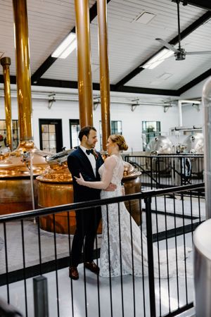 Newlyweds embracing in industrial-style brewery with copper brewing tanks.