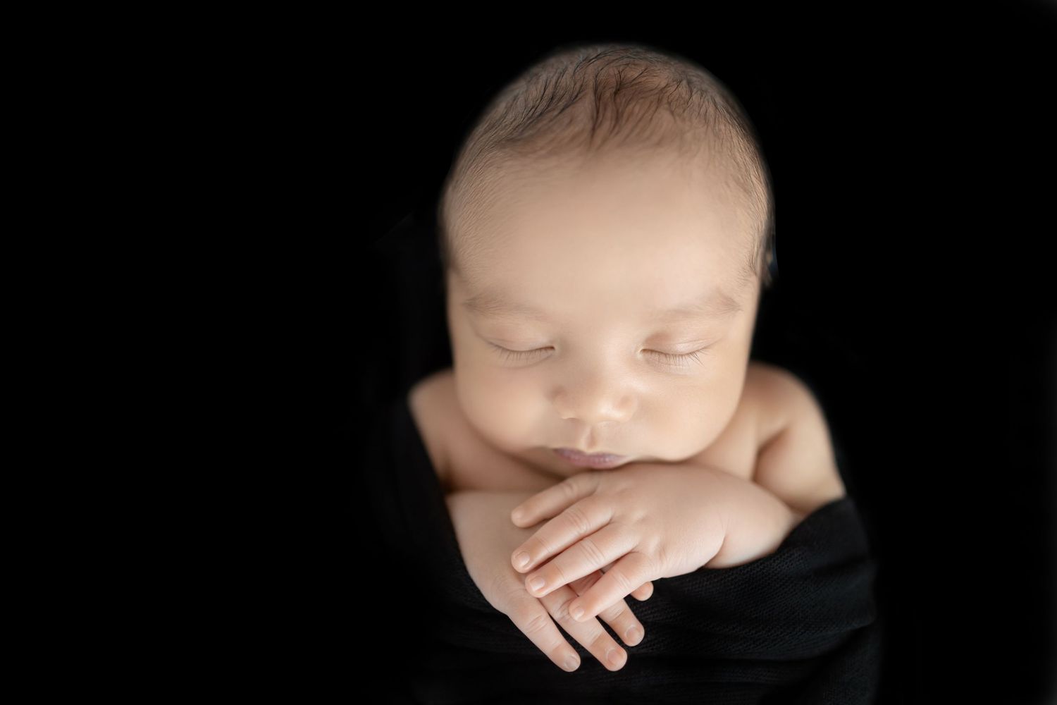 Peaceful sleeping newborn baby resting with hands folded against black background in artistic portrait.