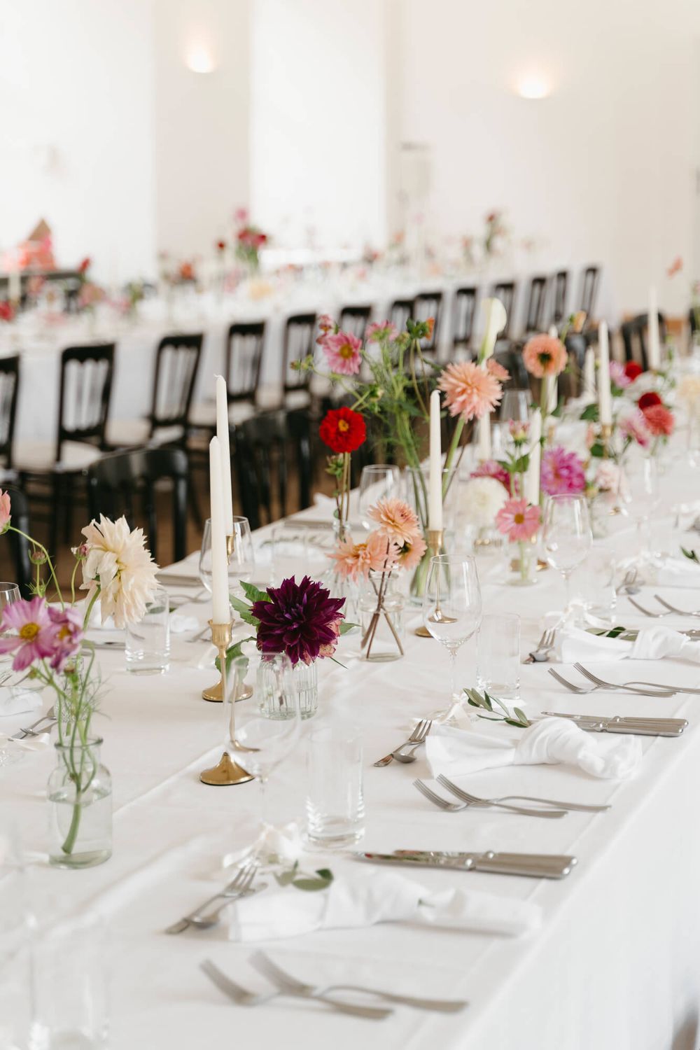 Elegant wedding reception table setting with white linens and colorful wildflower centerpieces in glass vases.