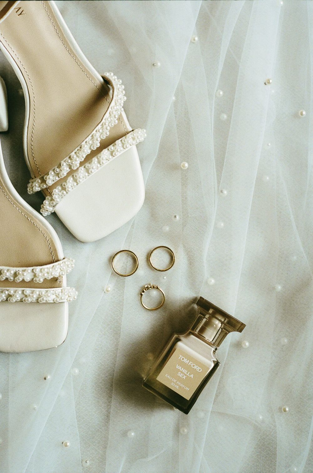 Wedding accessories including white heels, pearl details, gold rings and perfume bottle arranged on white tulle fabric.
