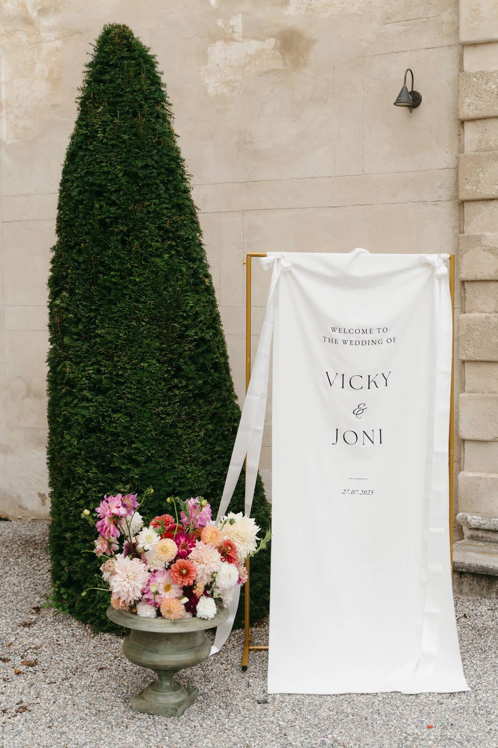 Elegant wedding signage and decor featuring topiary trees, floral arrangements, and white welcome banners against a stone wall.