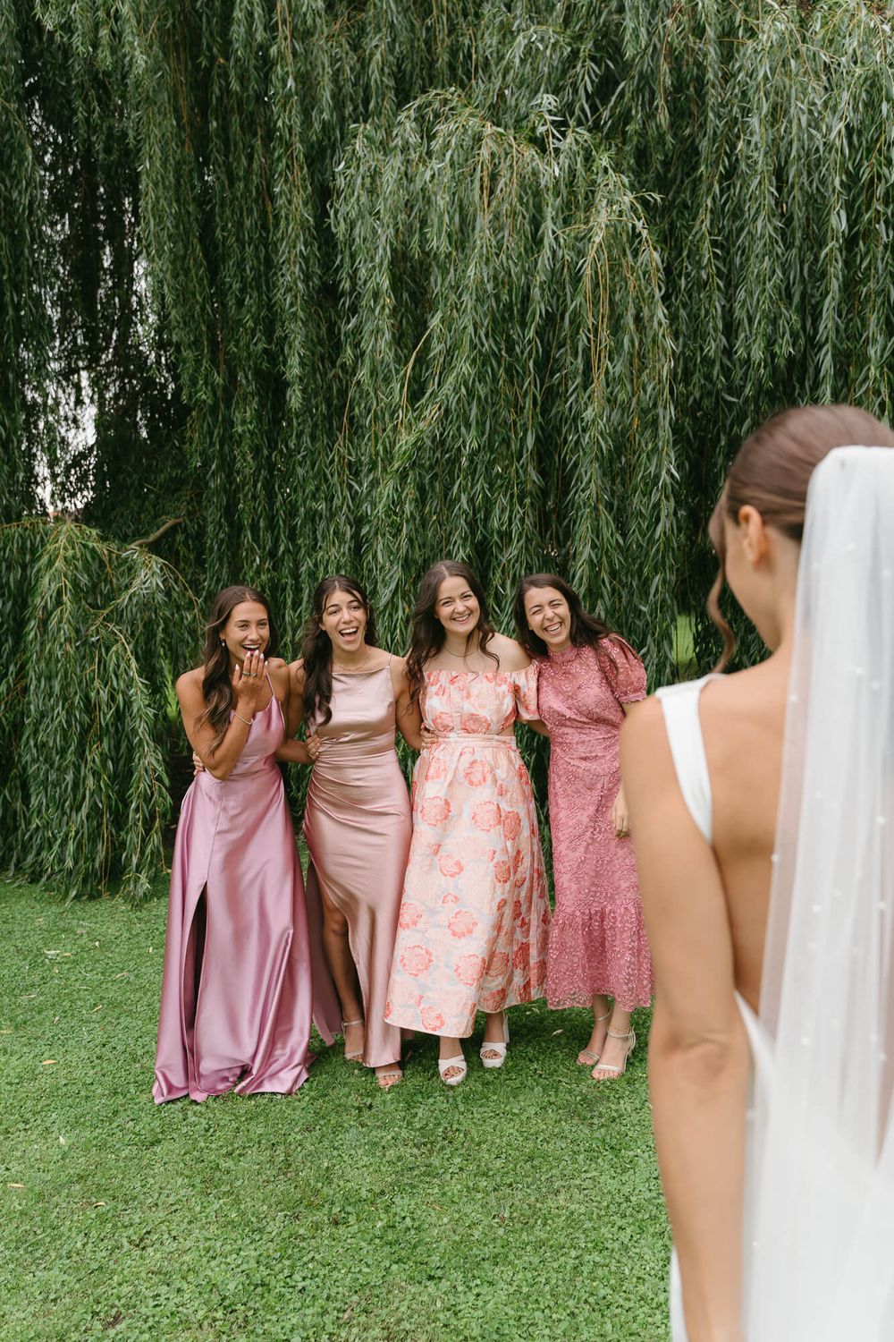 Bridesmaids in pink and coral dresses pose together under a weeping willow tree during an outdoor wedding celebration.