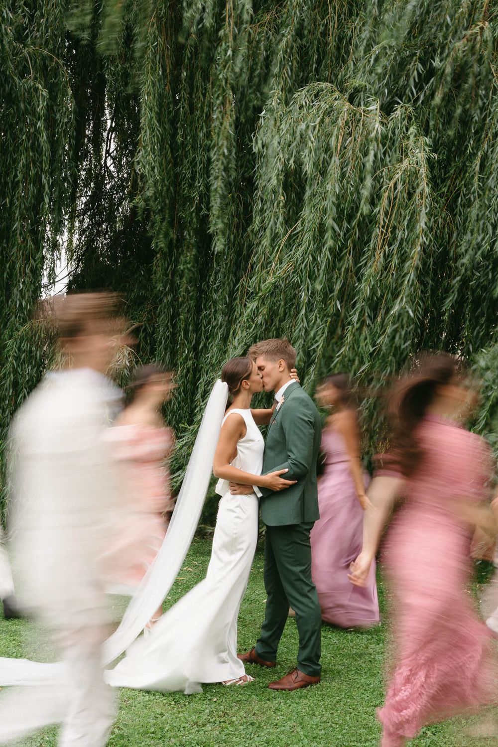 Motion blur sequence shows wedding party playfully running under drooping willow tree branches in a garden setting.