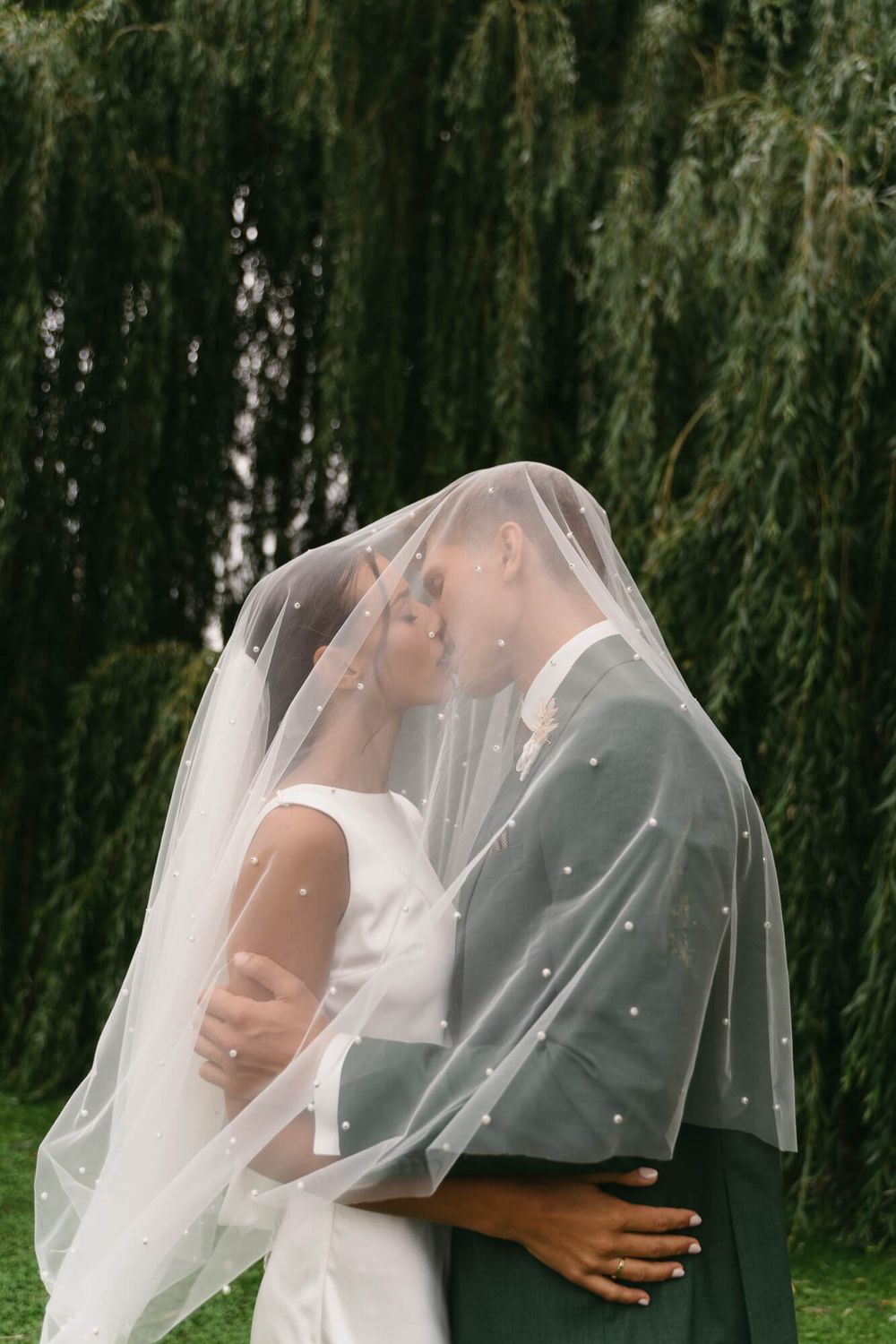 Wedding couple shares intimate moments under a flowing veil with willow trees creating a romantic backdrop.