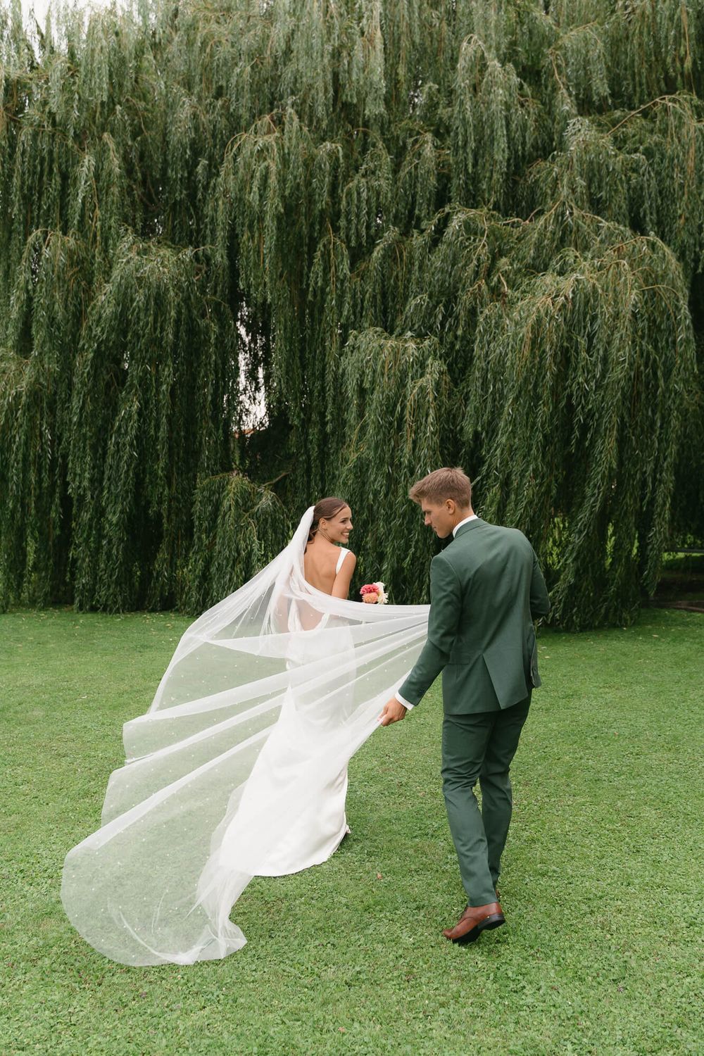 A bride and groom walk across a lawn with flowing veil against a backdrop of weeping willow trees.