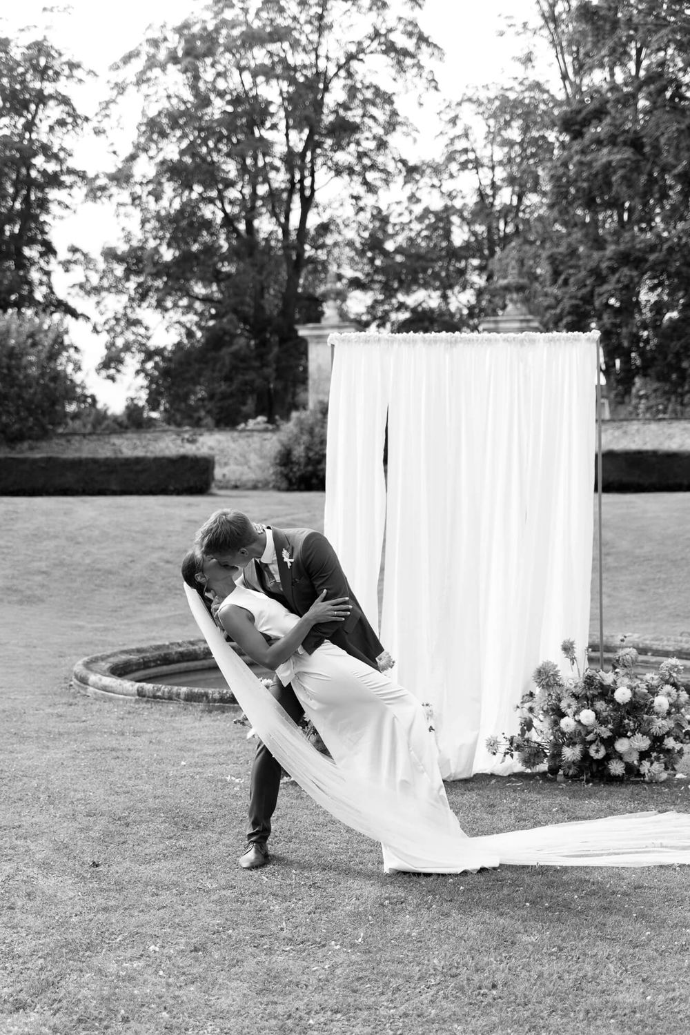 A sequence of four black and white photos showing a couple dancing with flowing white fabric on a garden lawn.