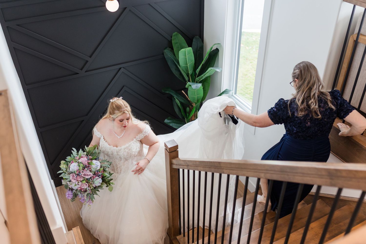 A bride in a flowing white gown ascends a wooden staircase while holding a pink and purple bouquet.