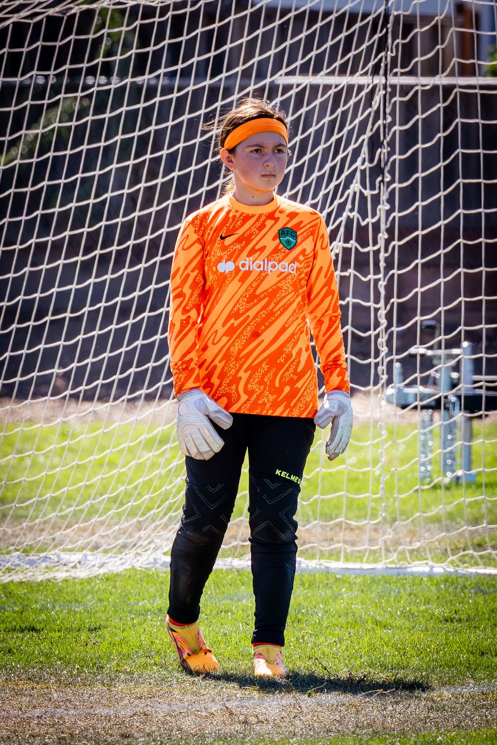 Soccer goalkeeper in bright orange jersey and black pants stands ready in front of goal net on sunny day.