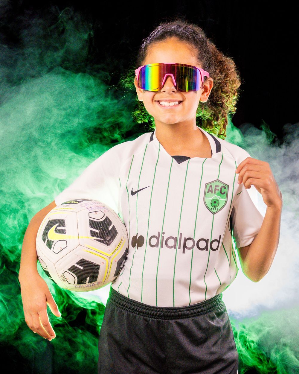Youth soccer player poses with ball wearing rainbow sunglasses and white uniform amid green smoke effects.