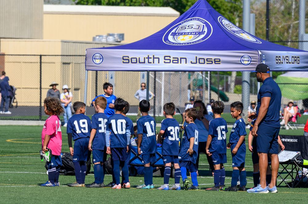 Youth soccer team kneeling together on a sports field with coaches and blue canopy tent.