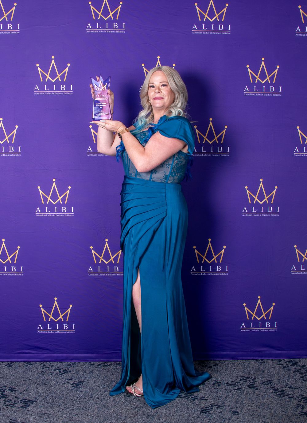 Award recipient poses with trophy against purple backdrop with crown logos wearing elegant teal evening gown.
