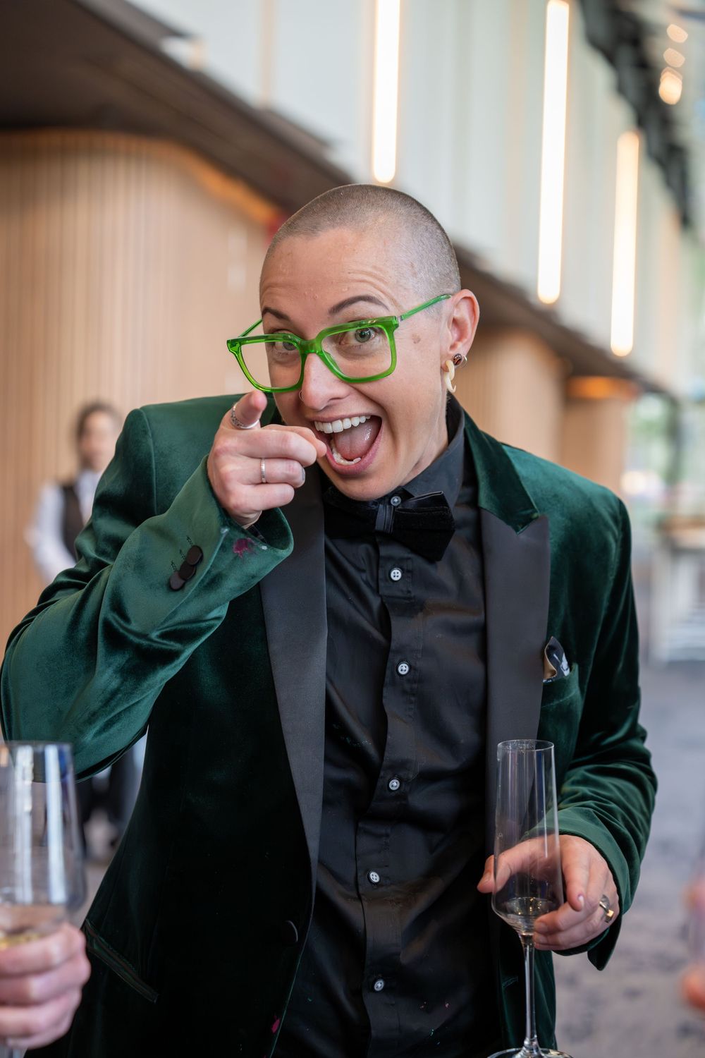 Person in green velvet blazer and glasses making playful expressions at formal event.