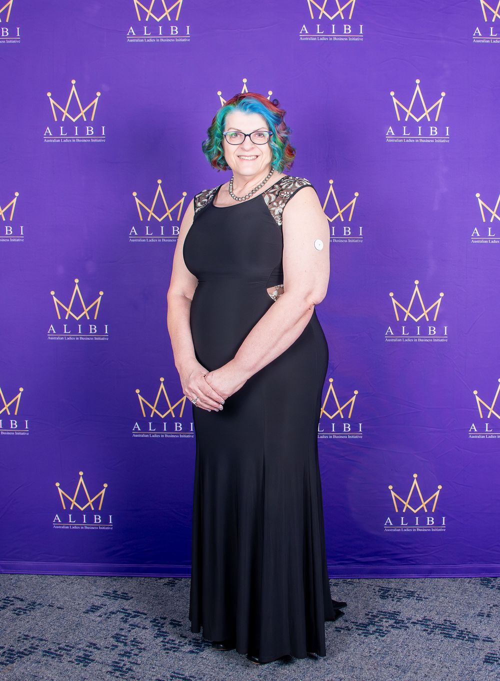 Formal event attendee in black gown poses against purple step-and-repeat wall with crown logos.