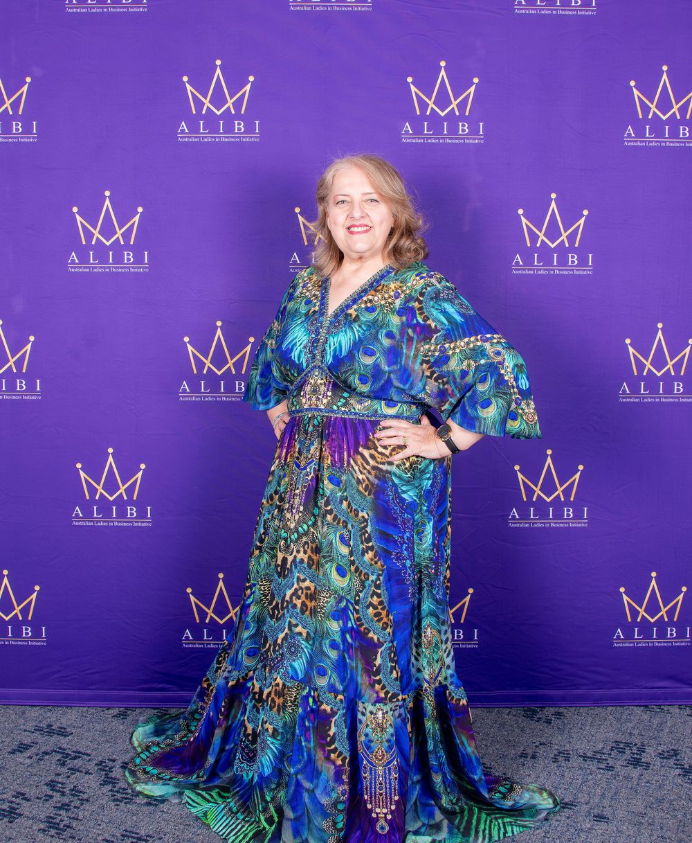 Guest wearing peacock-patterned blue and green gown poses at formal event with crown-decorated backdrop.