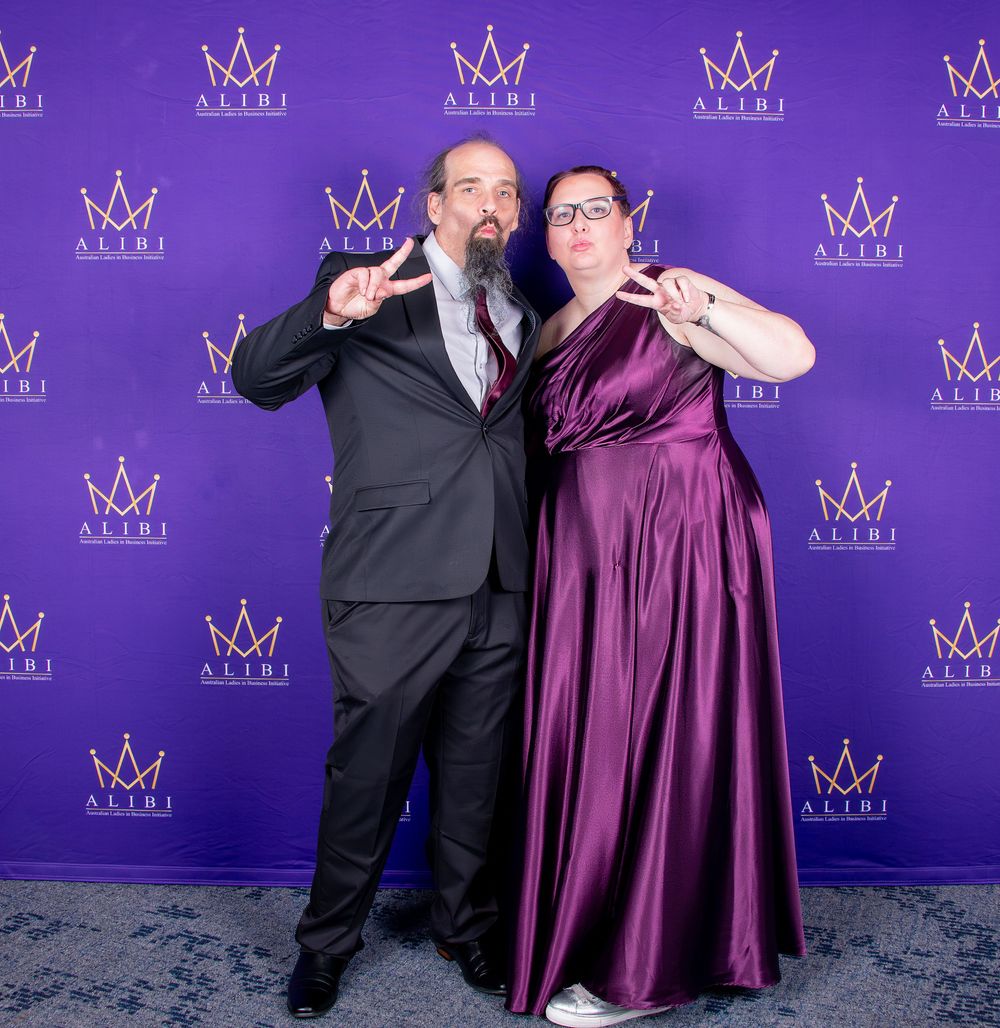 Couple in formal attire poses playfully against purple backdrop at awards ceremony.