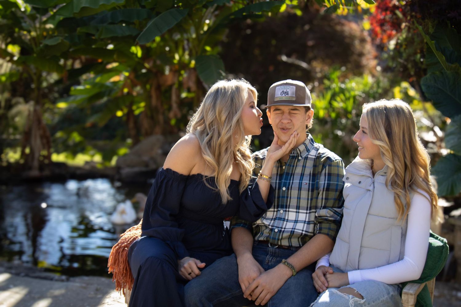 Three people sitting together and laughing on a bench in a sunny garden setting with a water feature in the background.