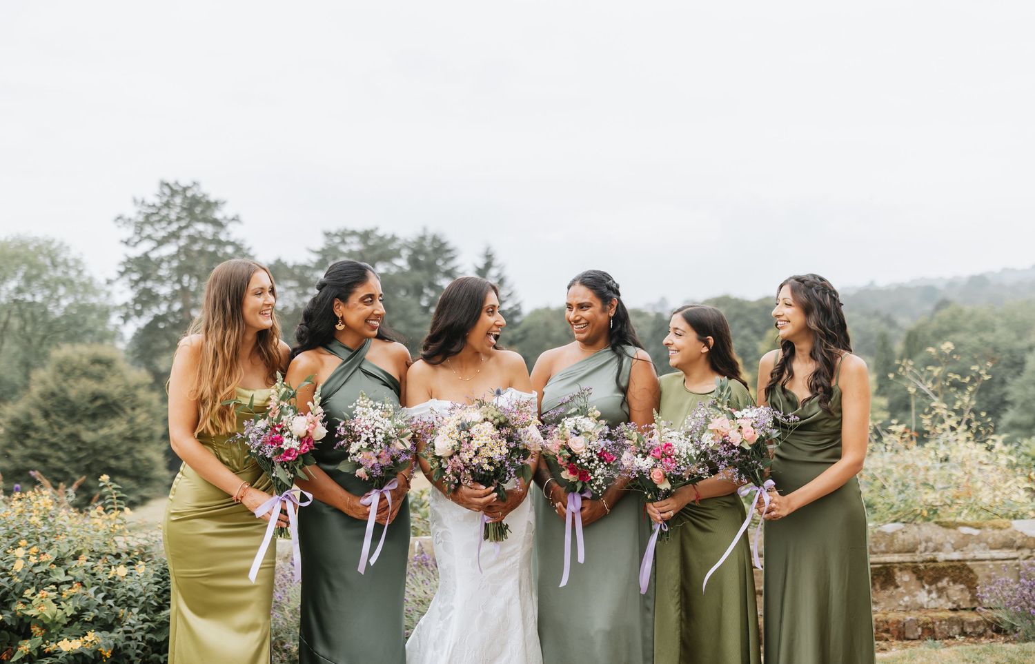 Wedding party in coordinated olive and sage green dresses pose together outdoors in natural setting.