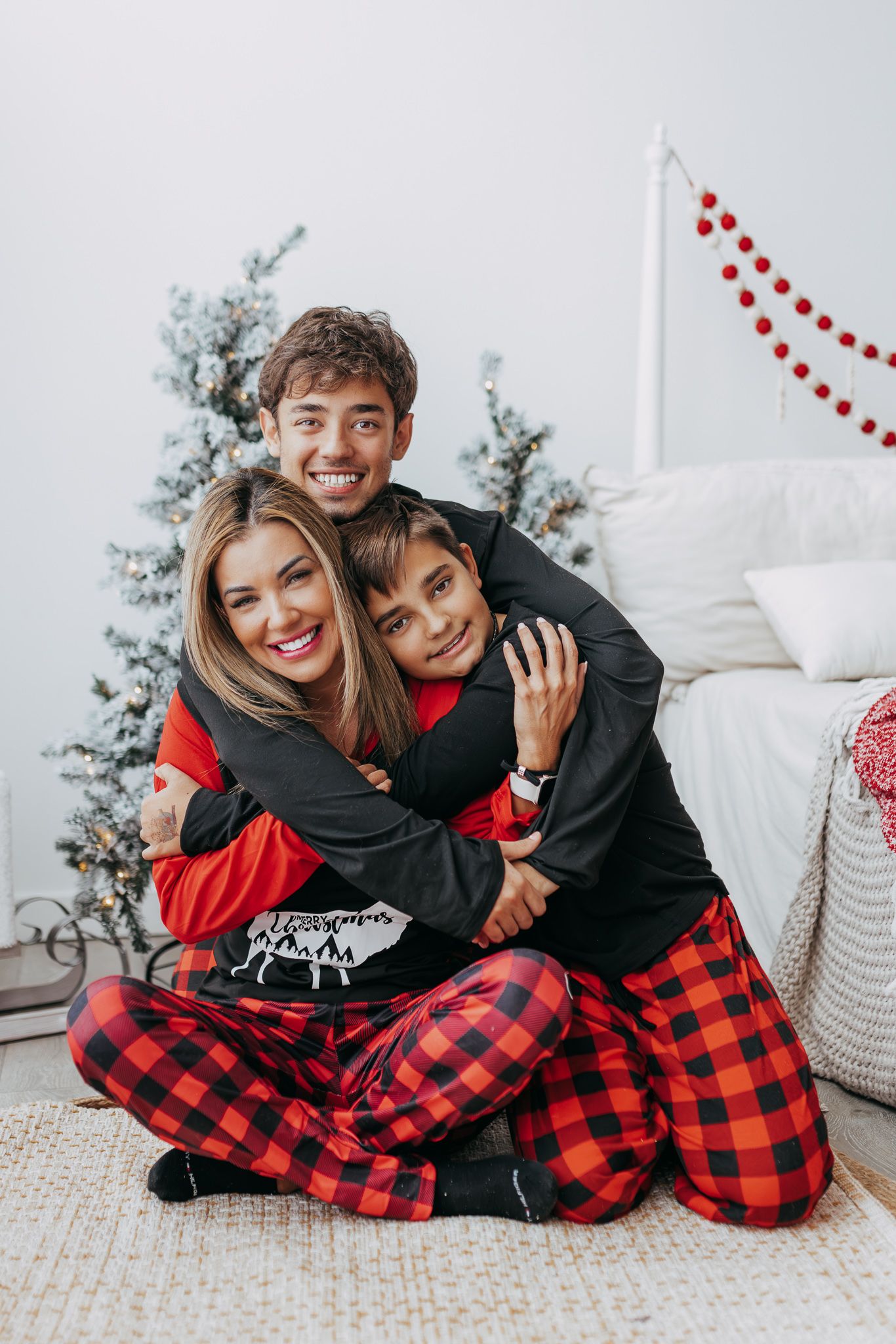 Family wearing matching buffalo plaid pajamas cuddle together on white bedding during holiday photo session.