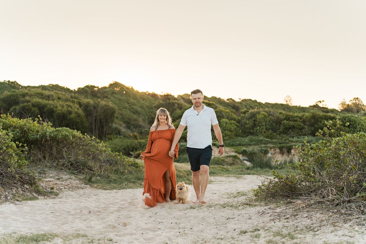 Couple walks together along a scenic coastal path through green vegetation at sunset with their dog during a maternity photoshoot in Sydney