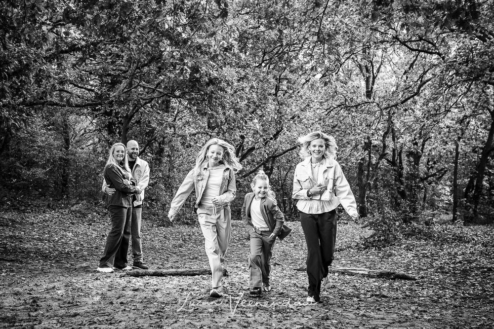 Dochters rennen lachend tussen de bomen terwijl hun ouders op de achtergrond toekijken.
