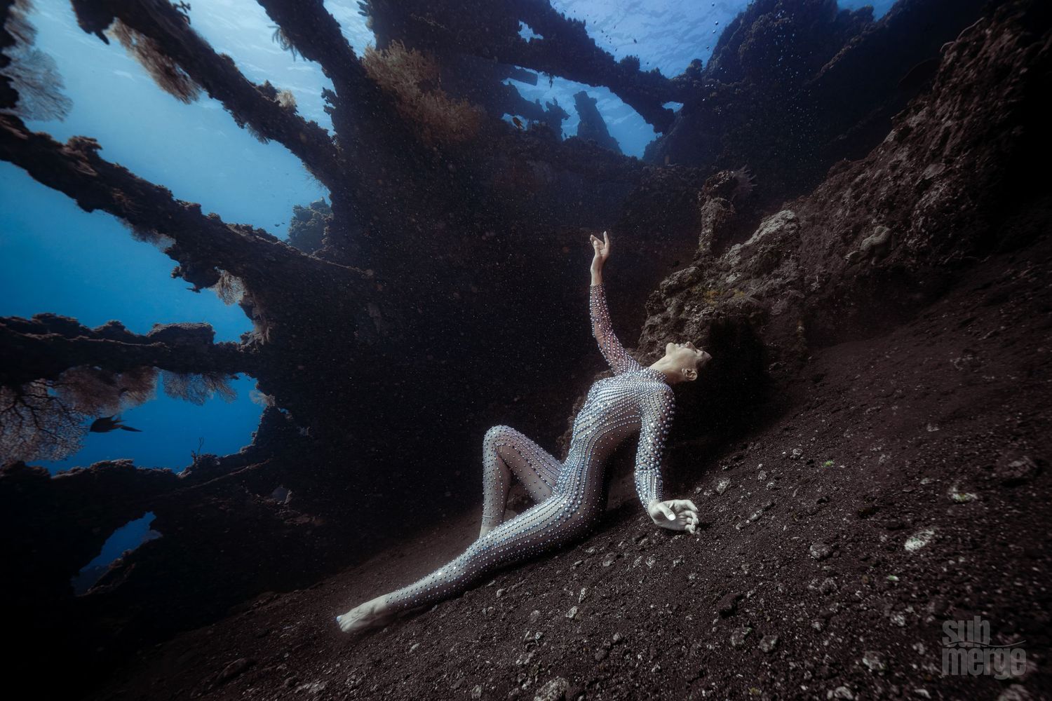 A figure in a flowing white dress poses gracefully against a dark rocky backdrop near water.