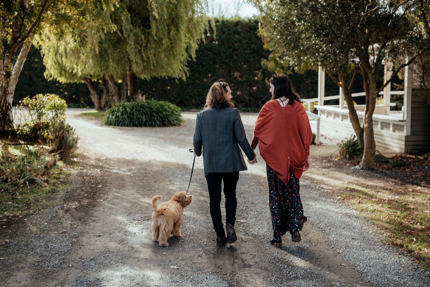Couple walk down a gravel path with a goldendoodle dog on a sunny autumn day at The Runholder Cottage by August and Vine