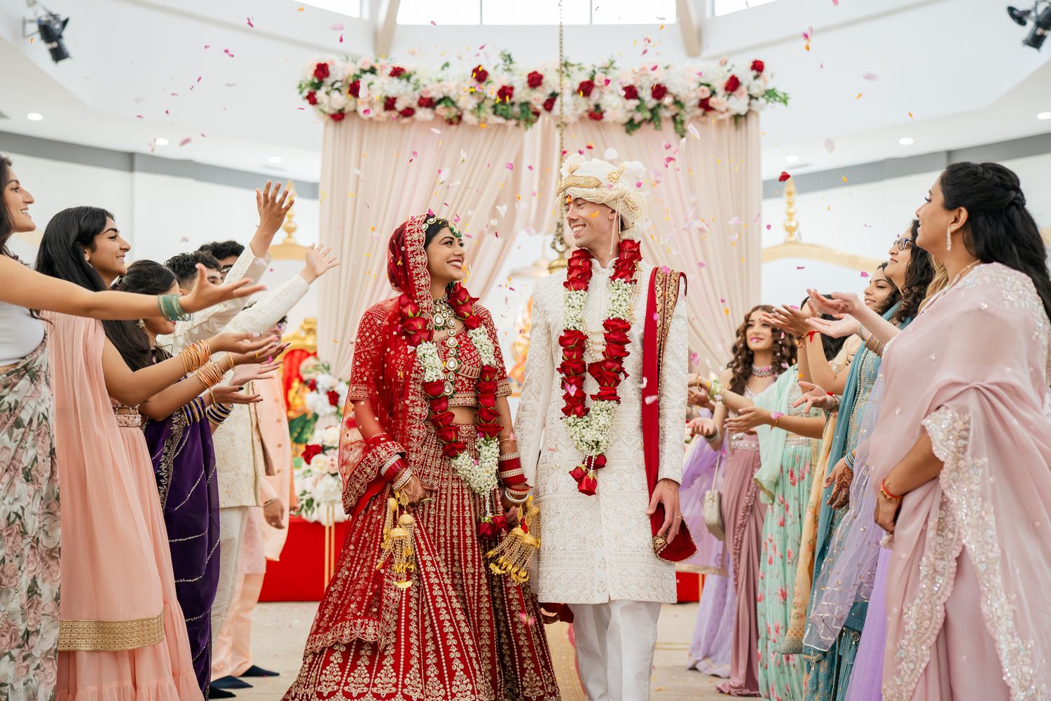 A couple in wedding attire runs joyfully together on a lush green lawn surrounded by evergreen trees.