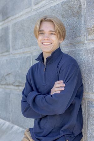 Smiling teen boy in navy pullover leaning against stone wall.