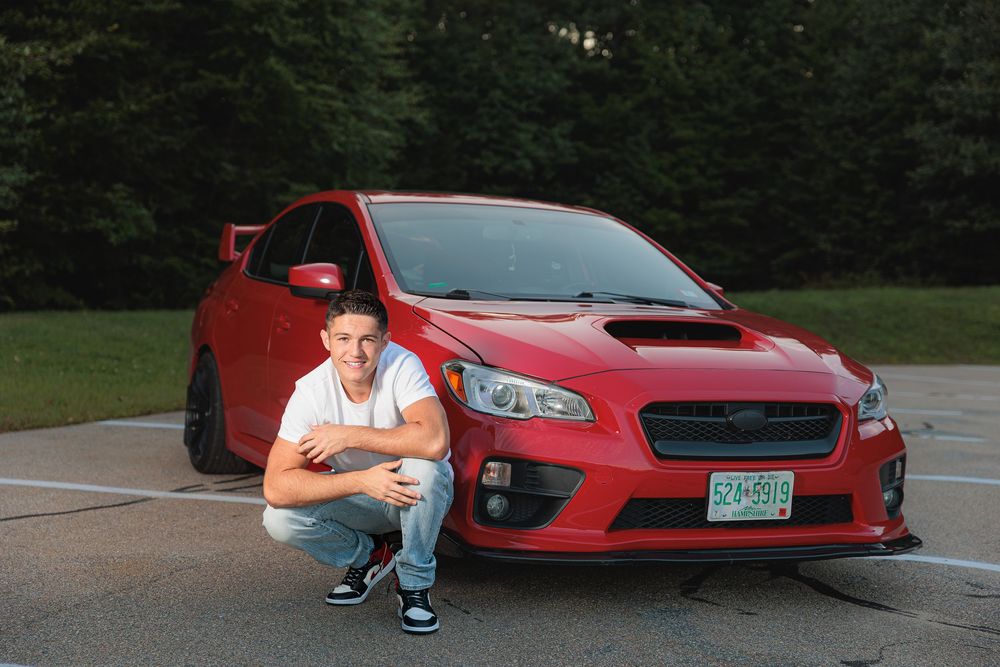 Young man crouches in front of red Subaru WRX STI in a parking lot surrounded by trees. Prospect Mountain High School Alton NH