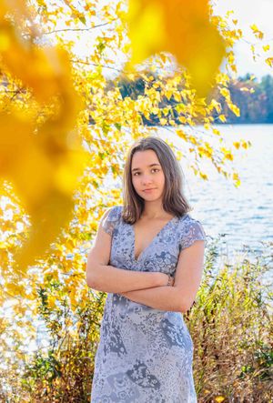 Teen girl in gray dress poses by autumn lake with golden leaves. Locke Lake Barnstead NH.