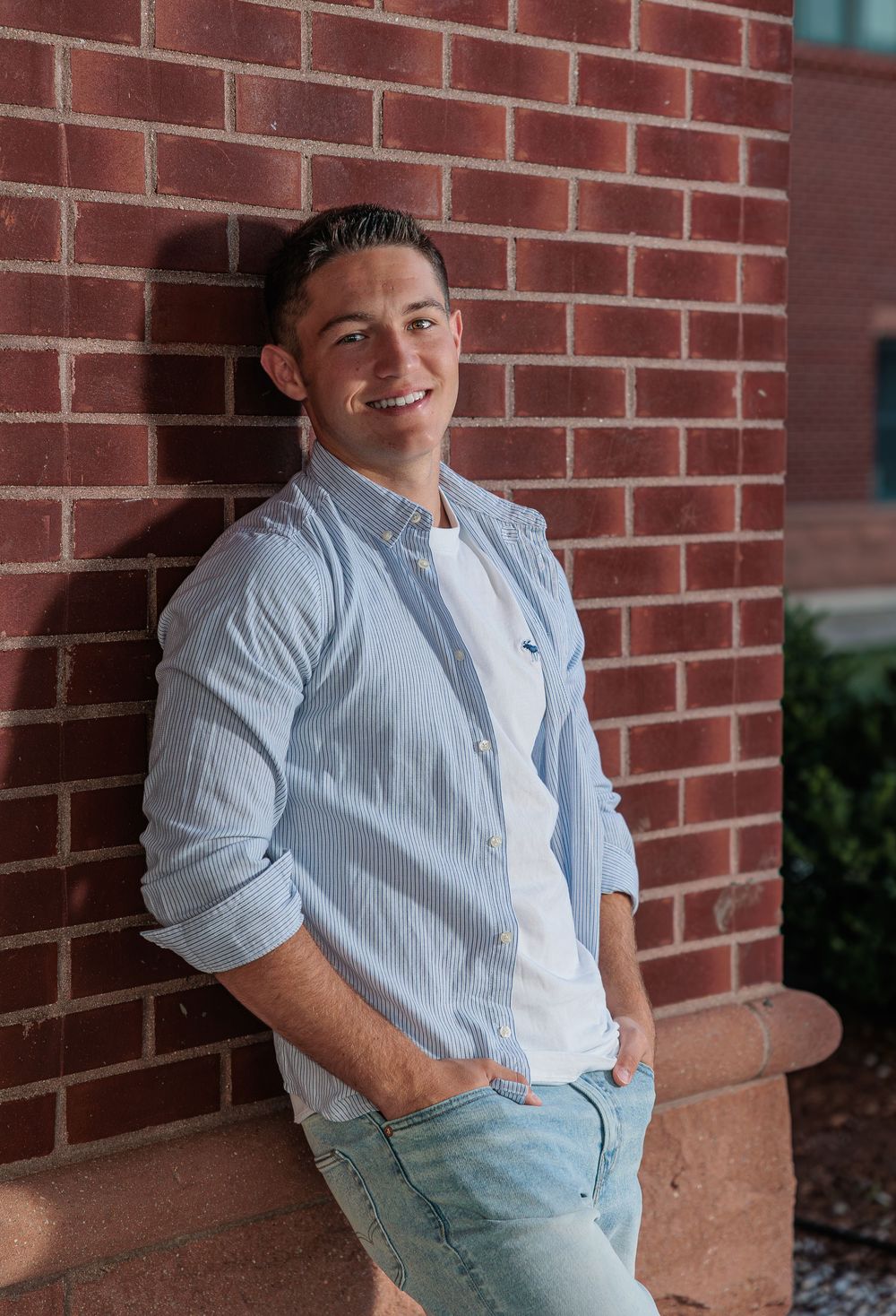 Smiling young man in light blue shirt leans against red brick wall with hands in pockets. Prospect Mountain High School Alton NH