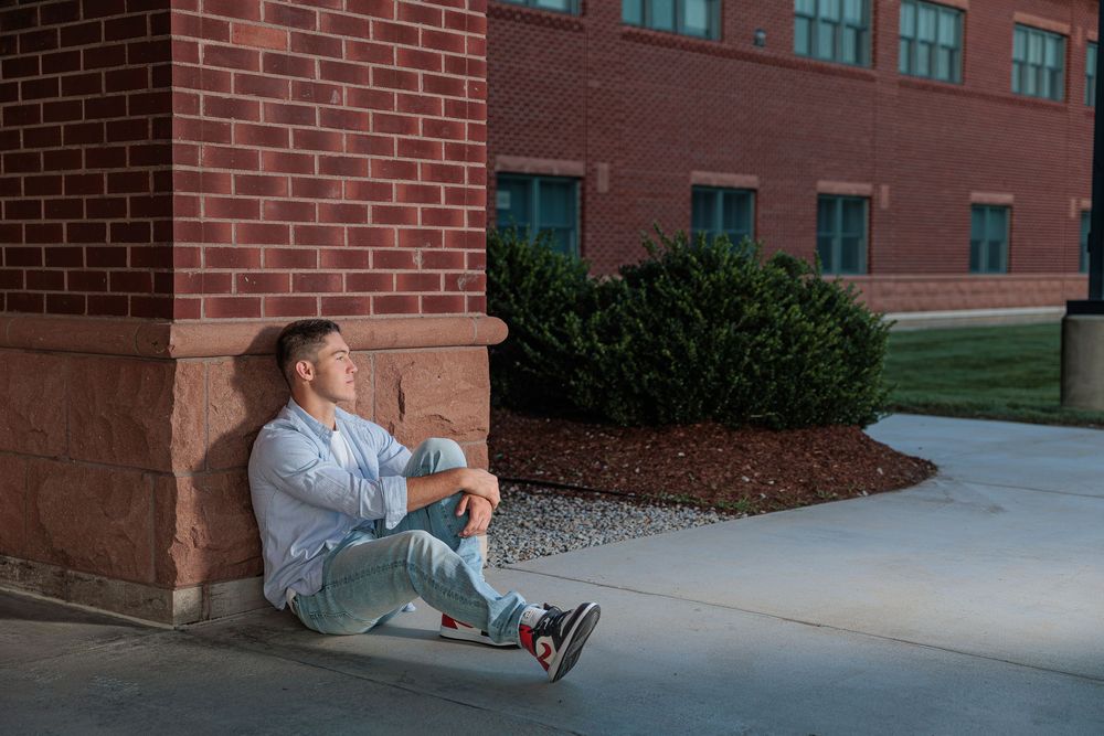 Young man in blue shirt sits relaxed against brick building corner on concrete sidewalk outdoors.