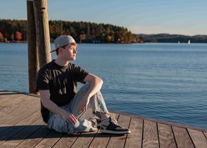 Young man in black t-shirt sitting on wooden dock overlooking calm lake in autumn. Cate Park Wolfeboro NH