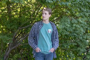 Young man in plaid shirt and green tee standing casually outdoors among lush green trees and foliage. Cate Park Wolfeboro NH