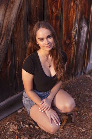Teen girl in black top crouching by rustic wooden barn wall. Cotton Valley Rail Trail, Wolfeboro NH.