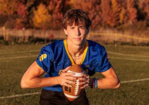 Teen boy in blue football jersey holding football on field with fall foliage background.