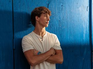 Young man in white t-shirt leaning against blue wooden wall with arms crossed.